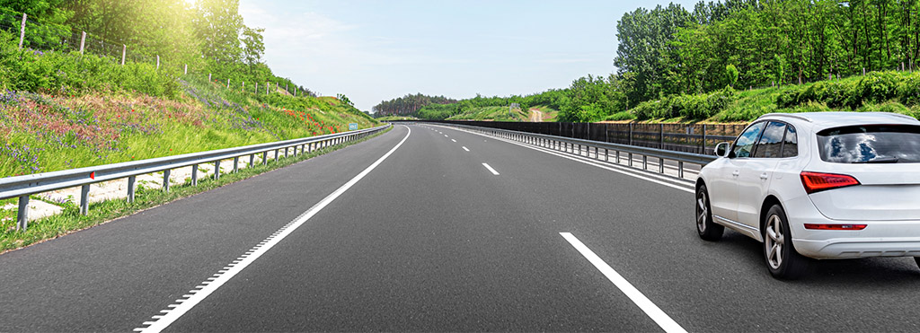 White SUV driving on a clear, empty highway surrounded by green trees - www.compassinsurancegroup.com